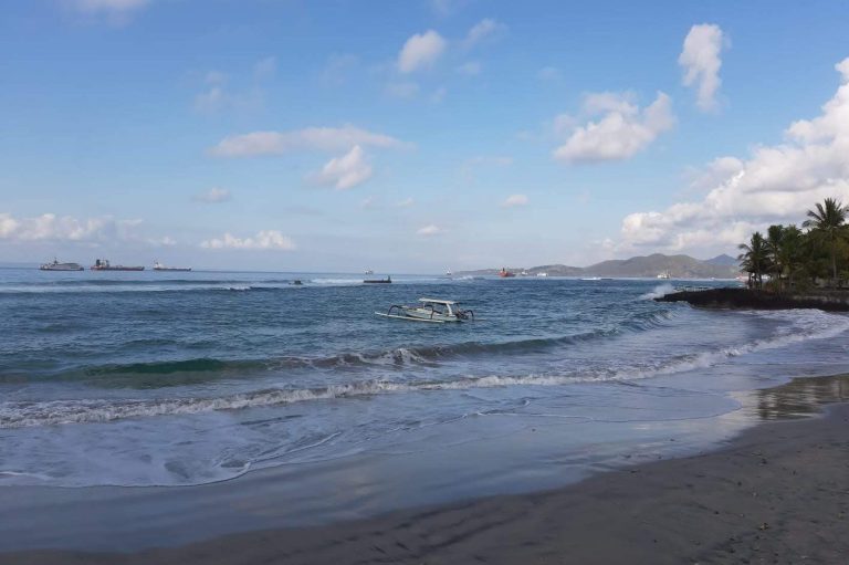 Strand mit sanften Wellen, Boot auf dem Wasser und grünen Bäumen im Hintergrund.