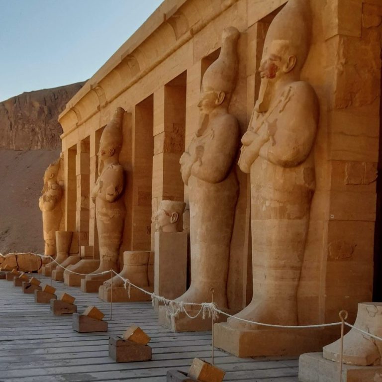 Drei antike Statuen stehen vor einer Tempelanlage in einer Wüstenlandschaft.