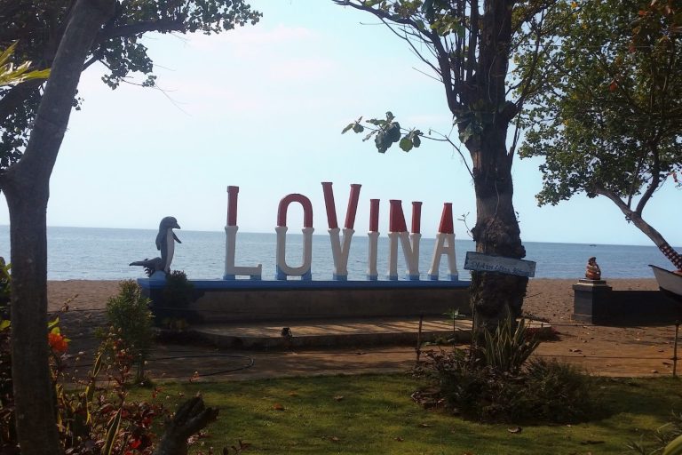 Bunte "Lovina"-Skulptur am Strand mit Palmen im Hintergrund.